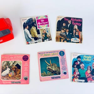May include: A red View-Master viewer sits next to five vintage View-Master reels. The reels feature images from "The Mod Squad," "The Addams Family," "Bobby the Bunny," "Battle of the Monsters," and "Family Affair."