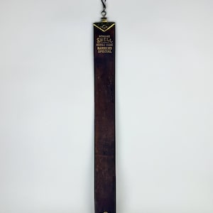 Puede incluir: Una correa de cuero vintage con el texto "GENUINE SHELL HORSE HIDE BARBERS SPECIAL". La correa es de color marrón oscuro con una parte superior de metal color latón y un lazo para colgar. La parte inferior de la correa tiene un emblema dorado y el texto "MADE IN U.S.A."