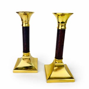 May include: A pair of candlestick holders featuring polished gold-tone brass bases and flared tops, connected by a central cylindrical dark brown wooden shaft. The square-shaped bases provide stability for these two matching metal and wood items.