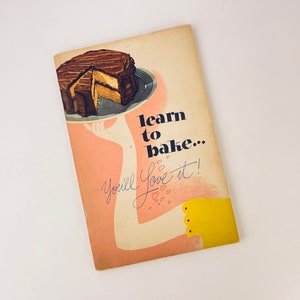 May include: A vintage cookbook cover with a pink background and a hand holding a plate with a chocolate cake. The text on the cover reads "learn to bake... You'll love it!"