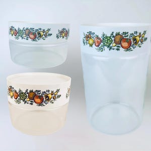 May include: Three clear, vintage-style plastic containers. Each container has a white band around the top with a colorful fruit and vegetable pattern. The containers vary in size, with one tall and two shorter, wider ones.