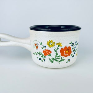 May include: White ceramic pot with a black lid and a floral design featuring orange, yellow, and blue flowers. The pot has a handle.