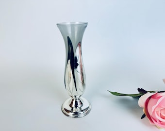 May include: A silver-colored vase with a flared top and a round base. The vase has a gray-tinted upper portion and decorative leaf-like designs. A pink rose is visible in the background.