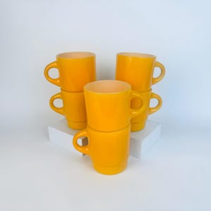 Puede incluir: Seis tazas de color amarillo brillante apiladas sobre una superficie blanca. Las tazas tienen forma redondeada con asa e interior de color más claro. Las tazas están dispuestas en dos pilas de tres.
