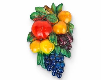 May include: A decorative wall plaque featuring a relief sculpture of assorted fruits including a bright red apple, yellow and orange peaches, dark purple plums, and a cluster of deep blue grapes, all surrounded by vibrant green leaves.