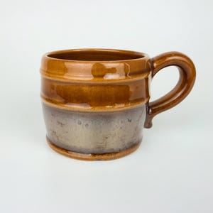 Op de afbeelding: Een keramische mok met een bruin en grijze glazuur. De mok heeft een afgeronde vatvorm met een handvat. De bovenkant en het handvat zijn glanzend bruin, terwijl het onderste deel een gedempt grijs is met bruine ondertonen.