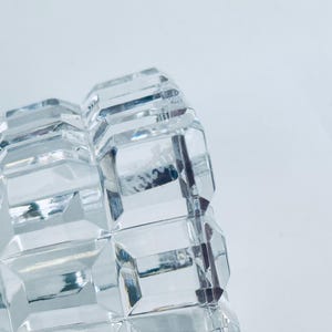 May include: Close-up of a clear, faceted glass object. The object has a repeating geometric pattern of rectangular shapes. The glass reflects light, creating a sparkling effect. The background is a plain white.