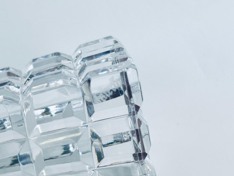 May include: Close-up of a clear, faceted glass object. The object has a repeating geometric pattern of rectangular shapes. The glass reflects light, creating a sparkling effect. The background is a plain white.