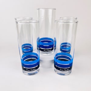 Puede incluir: Juego de cinco vasos altos transparentes. Cada vaso presenta un diseño de rayas horizontales azules y azul marino alrededor de la base. Las palabras "Pierre Cardin" están impresas en negro en la base de cada vaso.