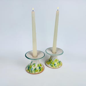 Peut inclure: Deux bougies coniques allumées, de couleur blanc cassé, dans des bougeoirs en céramique. Les bougeoirs sont blancs avec un motif floral vert et jaune. La base des bougeoirs est de couleur terre cuite. Les bougies sont centrées.
