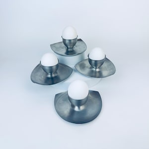 May include: Four metal egg cups with white eggs. Each cup is set on a silver-colored, leaf-shaped base. The arrangement is on a white surface, creating a clean, minimalist aesthetic.