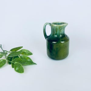 May include: A small, green ceramic jug with a handle and spout. The jug has a glossy finish with a gradient of dark and light green hues. A sprig of green leaves is to the left of the jug against a white background.