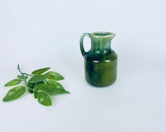 May include: A small, green ceramic pitcher with a handle and spout. The pitcher has a glossy finish with a gradient of dark and light green hues. A sprig of green leaves is to the left of the pitcher against a white background.