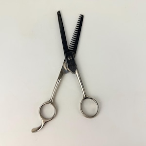 Könnte beinhalten: Professionelle Haarausdünnungsscheren mit schwarzer Klinge und silbernen Griffen. Die Schere hat eine kammartige Klinge zum Ausdünnen und eine Standardklinge. Die Griffe haben Fingerlöcher für eine einfache Handhabung.