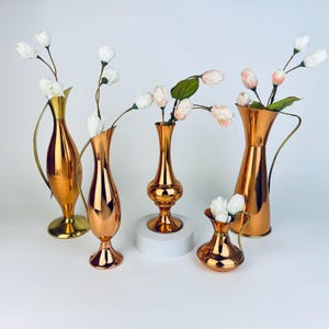 以下が含まれることがあります： さまざまな形とサイズの銅色の花瓶のコレクション。一部には取っ手が付いています。花瓶には白とピンクの花が活けられています。花瓶は白い表面に展示されています。