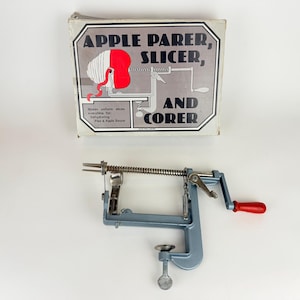 May include: An apple parer, slicer, and corer with a red handle and a silver clamp. The box above it reads "Apple Parer, Slicer, and Corer" with an illustration of an apple being peeled. The parer is made of metal.