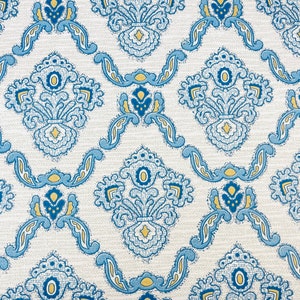 May include: Close-up of a patterned fabric with a repeating design. The pattern features blue and yellow floral motifs on a cream-colored background. The design is symmetrical and ornate, creating a vintage aesthetic.