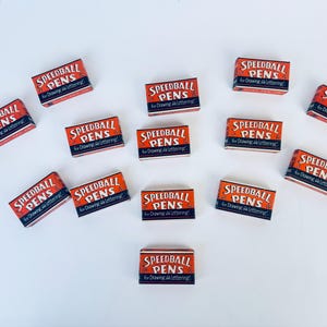 May include: A collection of 13 vintage Speedball pens for drawing and lettering. The boxes are orange with black lettering and are arranged in a heart shape on a white background.