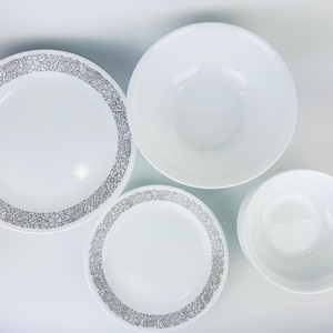 Puede incluir: Cuatro platos de cerámica blanca, dos con un patrón floral plateado alrededor del borde y dos lisos. Los platos están dispuestos sobre una superficie blanca.