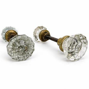 May include: Two clear, faceted glass door knobs with an octagonal shape and a textured surface. Each knob is attached to a circular brass-toned metal base and connected by a dark, square-shaped metal spindle that extends between the two pieces.