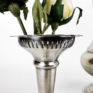 May include: A silver-toned metal vase with a flared, slotted rim holding artificial pink and yellow roses. Beside it stands a taller, smooth silver-toned metal vase with a rounded body and a wide base, both featuring a polished metallic finish.