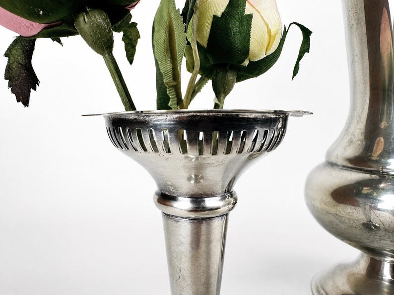 May include: A silver-toned metal vase with a flared, slotted rim holding artificial pink and yellow roses. Beside it stands a taller, smooth silver-toned metal vase with a rounded body and a wide base, both featuring a polished metallic finish.
