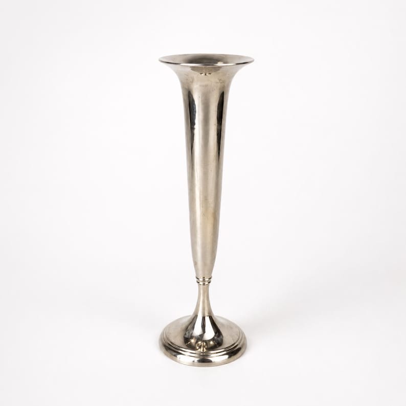 May include: A tall, slender silver-toned metal bud vase with a flared trumpet-shaped opening at the top. The vase features a smooth, polished surface that tapers down to a narrow neck, resting on a circular, stepped base for stability and balance.
