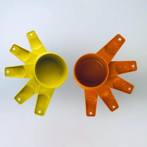 Może przedstawiać: Dwa zestawy miarek, jeden żółty i jeden pomarańczowy, ułożone na białej powierzchni. Każdy zestaw zawiera duży kubek z czterema mniejszymi kubkami, przypominającymi kształt gwiazdy. Kubki są wykonane z tworzywa sztucznego.