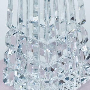 May include: Close-up of a clear, faceted crystal object. The object has a geometric design with stacked, square-like shapes at the base and a tapering, rectangular top. The crystal reflects light, creating a sparkling effect. The object is likely a decorative item.