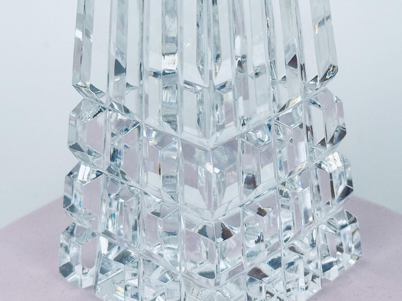 May include: Close-up of a clear, faceted crystal object. The object has a geometric design with stacked, square-like shapes at the base and a tapering, rectangular top. The crystal reflects light, creating a sparkling effect. The object is likely a decorative item.