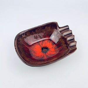 以下が含まれることがあります： 花柄のブラウンとオレンジのセラミック製灰皿。灰皿は角が丸みを帯びた長方形で、タバコ用のスロットが4つあります。