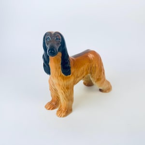 Puede incluir: Figura de cerámica de un perro afgano. El perro tiene un pelaje largo y fluido en tonos marrón y beige, con orejas negras y nariz negra. El perro está de pie sobre sus cuatro patas, con una apariencia detallada y realista.