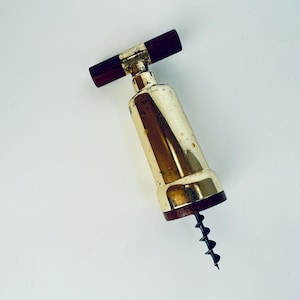 May include: A vintage wine bottle opener with a gold-coloured body and a dark brown wooden handle. The corkscrew is black and extends from the bottom. The opener is set against a white background.