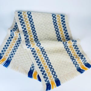 以下が含まれることがあります： 青と黄色のストライプが入った白い織りの毛布。毛布はフリンジの縁取りがされています。