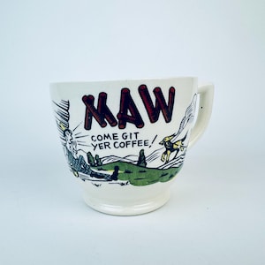 Puede incluir: Taza de café de cerámica blanca con una ilustración de dibujos animados de una persona y un perro. La taza tiene las palabras "KAW COME GIT YER COFFEE!" impresas en ella.