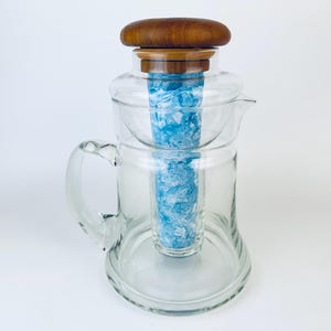May include: A glass pitcher with a wooden lid and a blue and white patterned filter inside. The pitcher has a handle and a base.