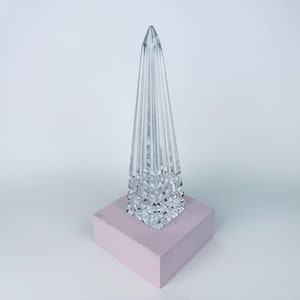 May include: A clear glass obelisk-shaped decorative object with vertical lines, set on a light pink square base. The top of the object is pointed. The base is a solid, opaque color.