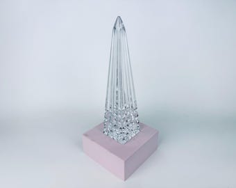May include: A clear glass obelisk-shaped decorative object with vertical lines, set on a light pink square base. The top of the object is pointed. The base is a solid, opaque color.
