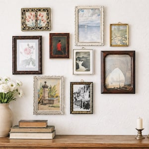 May include: A collection of framed art pieces, including paintings and prints, displayed on a white wall. The frames vary in style and color, from ornate gold to dark wood. A vase of white flowers, books, and candles sit on a wooden table below the artwork.