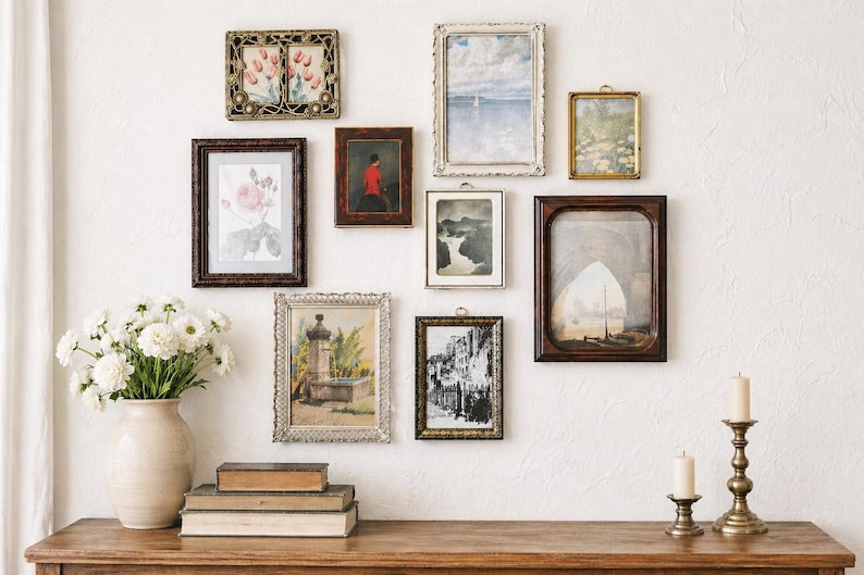 May include: A collection of framed art pieces, including paintings and prints, displayed on a white wall. The frames vary in style and color, from ornate gold to dark wood. A vase of white flowers, books, and candles sit on a wooden table below the artwork.