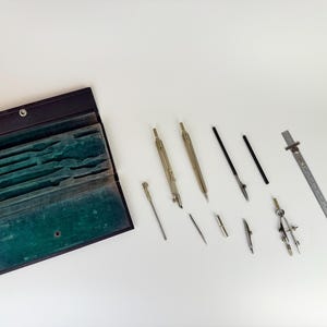 以下が含まれることがあります： 緑色のベルベットの内装が施された、開いた黒革ケース。さまざまな製図ツールが入っています。複数のコンパス、ディバイダー、定規が白い表面に配置され、精密機器のコレクションを紹介しています。