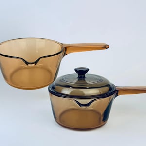 May include: Two amber-colored glass saucepans with integrated handles. One saucepan has a lid with a black knob. Both pans have a pouring spout. The cookware is made of transparent glass.