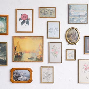 May include: A collection of framed art pieces, including landscape paintings, floral prints, and architectural sketches, arranged on a white wall. The frames vary in shape and size, with gold and wood finishes.