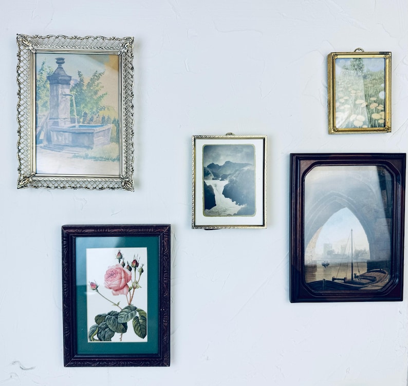 May include: A collection of framed art pieces, including a landscape painting of a fountain, a botanical print of a rose, and a vintage photograph of a waterfall. The frames vary in style, from ornate gold to dark wood.