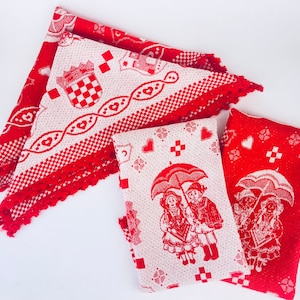 Può includere: Tre tovagliette all'americana in tessuto rosso e bianco intrecciato con un bordo all'uncinetto. Le tovagliette presentano un motivo ripetuto di cuori, una corona e un design a scacchi. Due delle tovagliette presentano una scena di due bambini che tengono degli ombrelli.