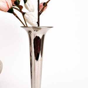 May include: A tall, slender, polished silver-toned metal vase with a flared trumpet-shaped opening. The vase has a smooth, reflective surface and contains several artificial flower stems with pale pink and white buds on brown branches.