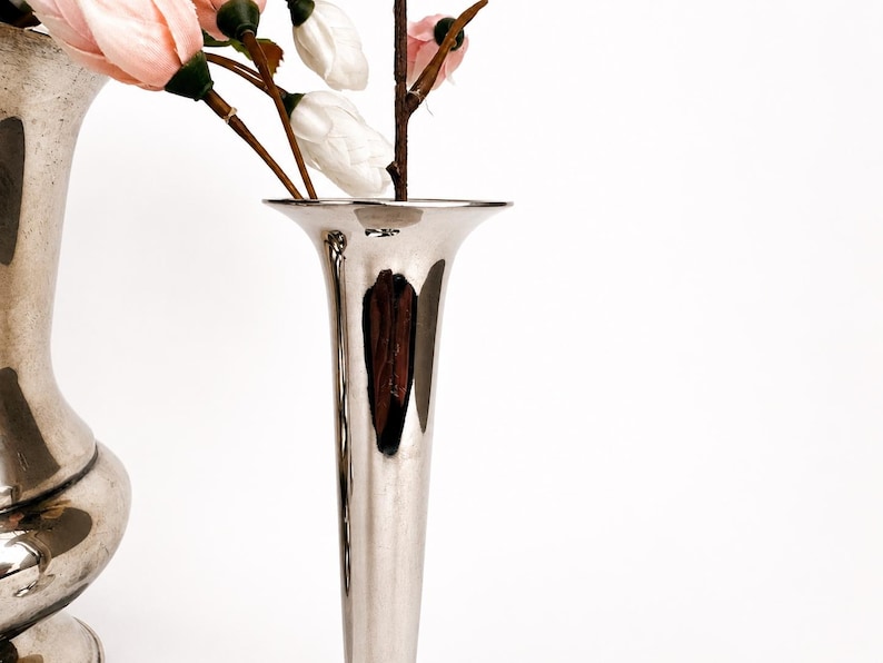 May include: A tall, slender, polished silver-toned metal vase with a flared trumpet-shaped opening. The vase has a smooth, reflective surface and contains several artificial flower stems with pale pink and white buds on brown branches.