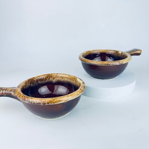 May include: Two brown and tan glazed ceramic bowls with handles. The bowls are small and round with a brown glaze and a tan rim.