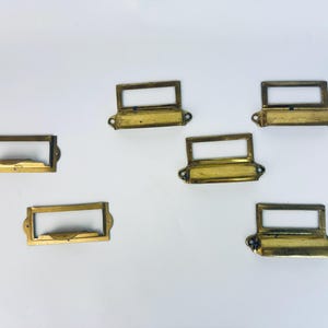 Puede incluir: Seis portatarjetas de latón vintage. Cada uno tiene un marco rectangular y bordes redondeados, con una abertura rectangular para etiquetas. Se ven pequeños agujeros para tornillos. El latón tiene una pátina cálida y envejecida.