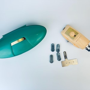 Op de afbeelding: Een tealblauwe plastic koffer met een gouden accent en een beige elektrisch scheerapparaat met een gele knop. Er zijn vijf kleine blauwe plastic stukjes en een metalen stuk met twee gaten.